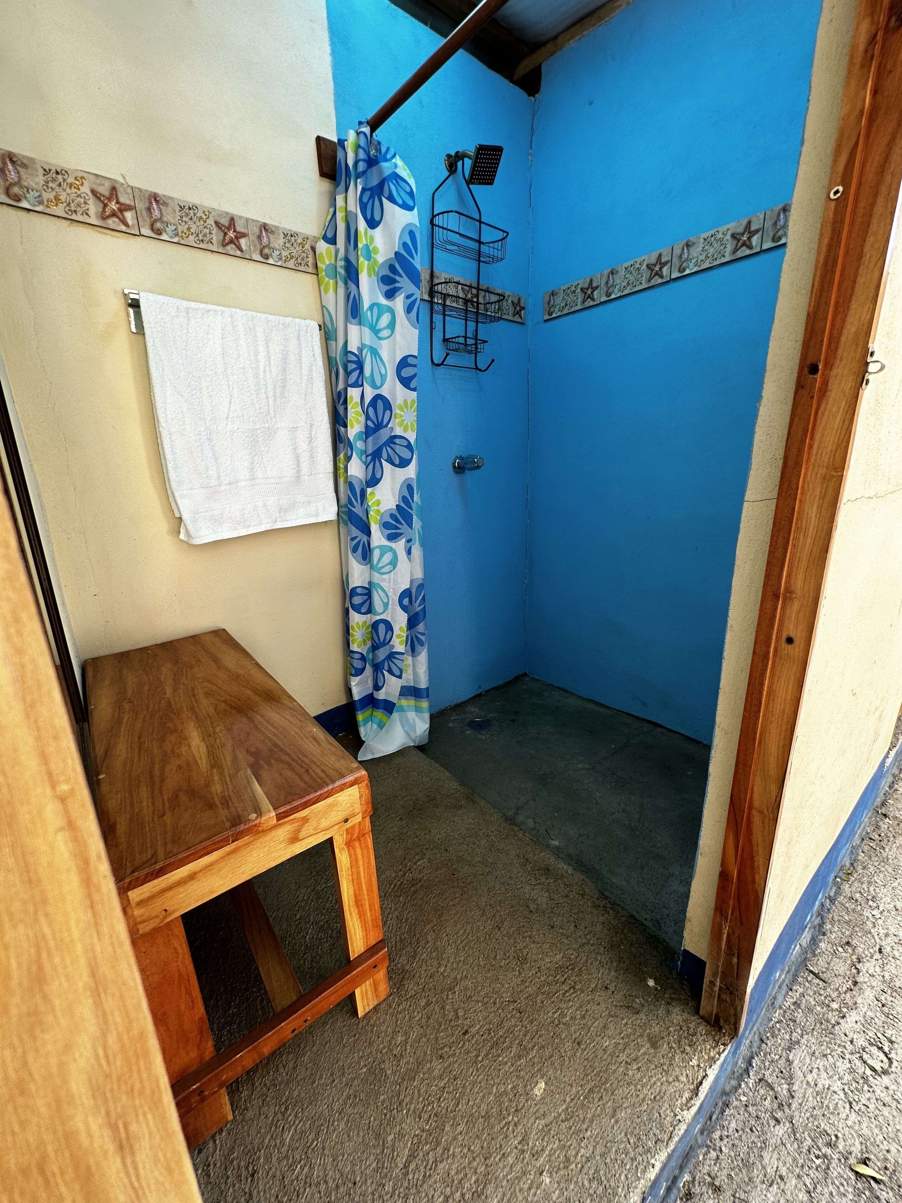 Une petite salle de bain fermée, équipée d’une douche, d’un tabouret en bois et d’une serviette accrochée au mur.