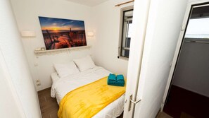 Ein kleines, ordentlich eingerichtetes Schlafzimmer mit einem Bett, einem Fenster, einem Sonnenuntergangsbild und einer Tür zu einem anderen Raum.