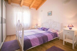 Un dormitorio con una cama, dos mesitas de noche, un ventanal con cortinas, techo de madera y un cuadro colgado en la pared.
