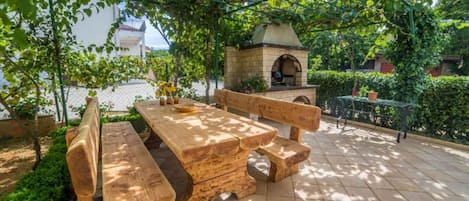An outdoor patio with a wooden table and benches, a brick oven, and a pergola with climbing plants.