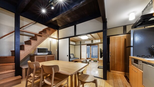 A modern kitchen with wooden flooring, a dining area with a wooden table and chairs, a built-in stove, and a kitchen island. - Shiki Homes Machiya Mei (Kyoto)