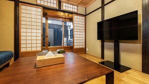 A traditional Japanese room with tatami flooring, a low wooden table, a futon bed, sliding shoji doors, and a television. - Shiki Homes Machiya Mei (Kyoto)