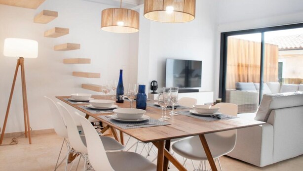 Un comedor moderno con una mesa de madera dispuesta para seis comensales, sillas blancas y dos lámparas de madera colgantes.