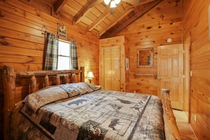 A wooden cabin bedroom with a bed, a window with plaid curtains, a wooden ceiling, and a door.