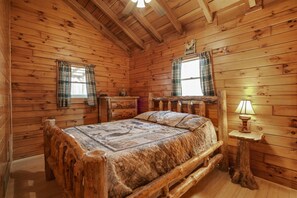 A wooden cabin bedroom with a bed, two windows with plaid curtains, a nightstand with a lamp, and a wooden headboard. - Moonlight Ridge Cabin | Hocking Hills, 200 Acres (South Bloomingville)