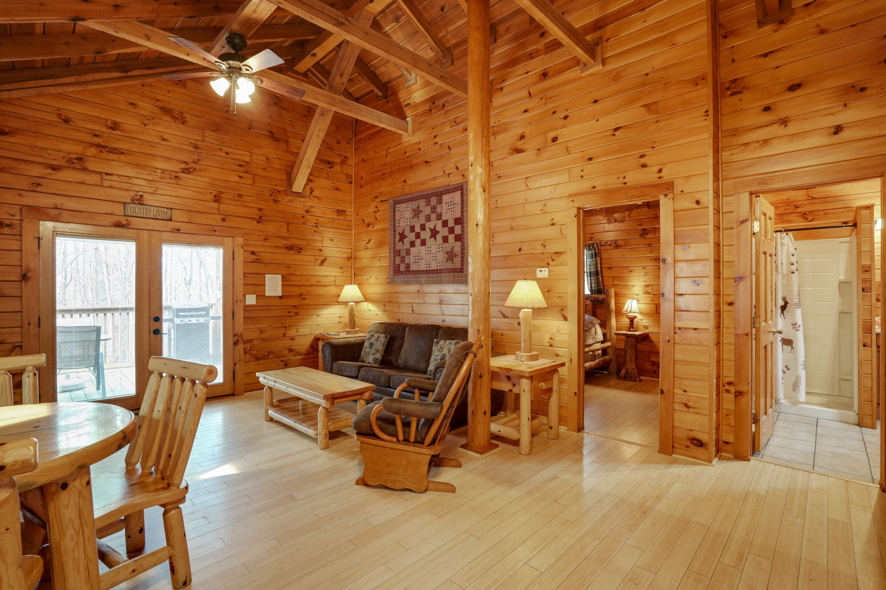 L’intérieur d’une cabane en rondins, avec un plancher en bois, un canapé, un fauteuil à bascule, une table, un ventilateur de plafond et une salle de bain visible à travers une porte ouverte.