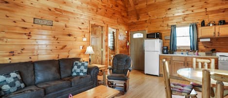 A cozy log cabin interior with a sofa, armchair, dining table, and kitchen area.