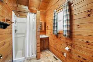A wooden cabin bathroom with a shower, toilet, sink, and a window with curtains. - Sunset Ridge Cabin | 200 Acres, Pets, Hot Tub (South Bloomingville)