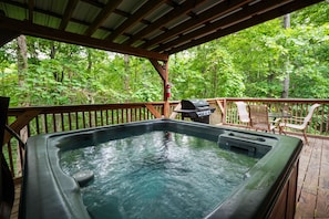 A hot tub on a wooden deck with a grill and chairs, surrounded by lush greenery. - Sunset Ridge Cabin | 200 Acres, Pets, Hot Tub (South Bloomingville)