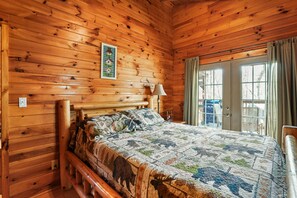 A wooden cabin bedroom with a bed, a nightstand, a lamp, and a window with curtains. - Sunset Ridge Cabin | 200 Acres, Pets, Hot Tub (South Bloomingville)
