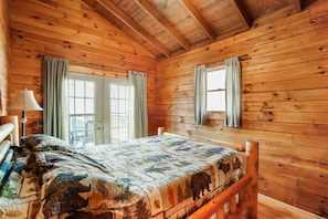 A wooden cabin bedroom with a bed, curtains, a window, and a lamp. - Sunset Ridge Cabin | 200 Acres, Pets, Hot Tub (South Bloomingville)