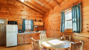 A wooden cabin interior with a kitchen, dining area, and a window with curtains. - Sunset Ridge Cabin | 200 Acres, Pets, Hot Tub (South Bloomingville)