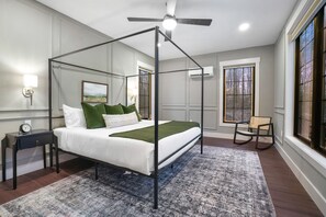 Una cama con dosel con ropa de cama blanca y verde, suelo de madera oscura, alfombra estampada, mesita de noche con un reloj, una silla y una ventana con vista a árboles.
