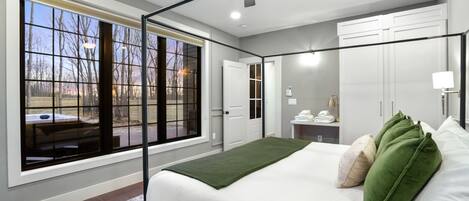 A bedroom with a large window, a bed with green and beige pillows, a ceiling fan, and a nightstand with a lamp.