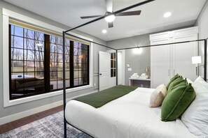 Un dormitorio con un ventanal grande, una cama con almohadas verdes y blancas, un ventilador de techo y una mesita de noche con lámpara.