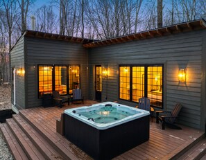 Une cabane en bois dotée d’un bain à remous sur la terrasse, de grandes fenêtres et d’un espace salon extérieur.