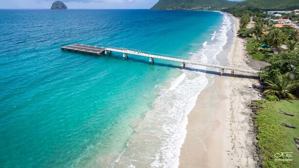 Paesaggio costiero con un molo che si protende in acque turchesi, una spiaggia con vista chiara su una montagna sullo sfondo e un piccolo paese con edifici e palme.