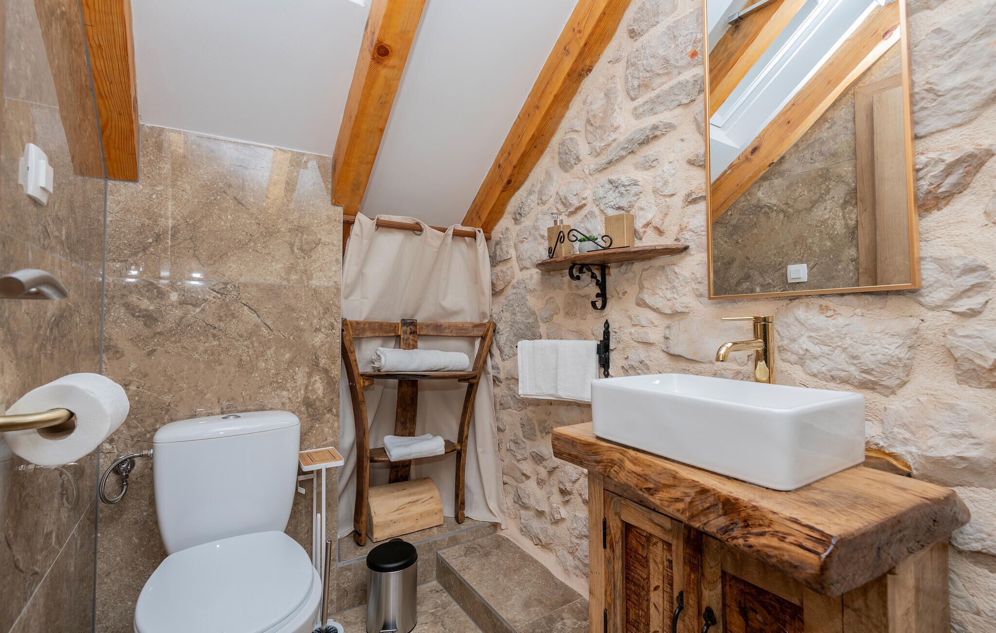 Une salle de bain avec un mur en pierre, des poutres en bois, un lavabo blanc et des toilettes.