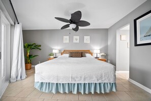 Un dormitorio con una cama grande, ventilador de techo, lámparas de noche, planta en maceta y cuadros en la pared.