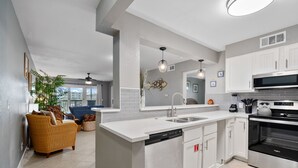 A modern kitchen with white cabinetry, a built-in dishwasher, a microwave, and a stove. There is a dining area with wicker furniture and a large window. - La Mirage Condo 214 Beachfront Resort Condominiums with Private Boardwalk (Port Aransas)