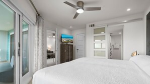 A bedroom with a large bed, a ceiling fan, a TV, and a sliding glass door leading to a balcony.