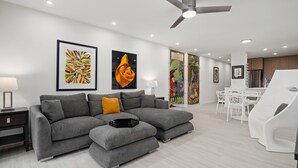 A modern living room with a grey sectional sofa, a small ottoman, and a dining area with white chairs.