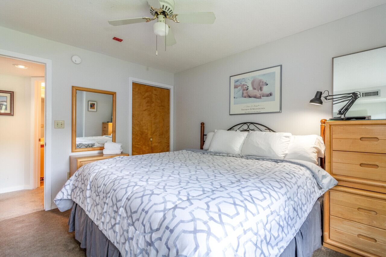 Een slaapkamer met een groot bed, een houten dressoir, een plafondventilator, een ingelijst schilderij aan de muur en een spiegel boven het dressoir.