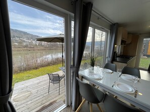 Un coin repas avec une table dressée pour deux personnes, donnant sur un lac et un paysage vallonné.