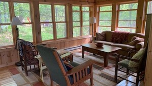 A cozy cabin interior with wooden walls, large windows, a sofa, chairs, a table, and a lamp.