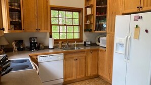 A kitchen with wooden cabinets, a white refrigerator, a dishwasher, a stove, and a window with a view of greenery.