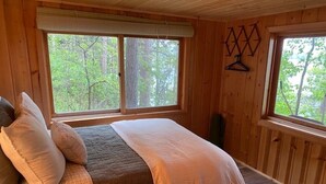 A wooden cabin bedroom with a bed, pillows, and two windows offering a view of the forest.