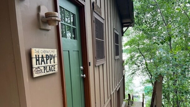 A cabin with a green door, a welcome mat, and a sign that reads "The Cabin is My Happy Place."