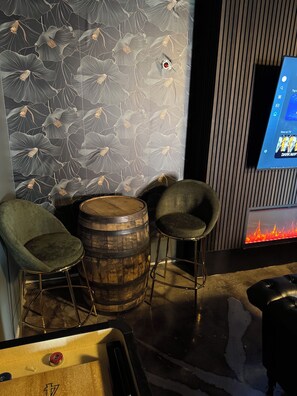 A bar area with two stools, a wooden barrel, and a flat-screen TV displaying a fire.