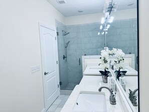 Features a sleek countertop, single sink, and large mirror, with a shower area and marble patterned walls.