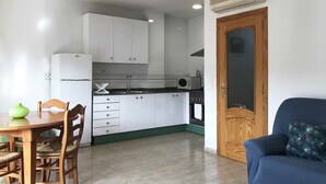 Eine kompakte Küche mit weißen Schränken, einer Mikrowelle und einem Herd. Im angrenzenden Wohnbereich befindet sich ein Esstisch mit Stühlen sowie ein blaues Sofa.