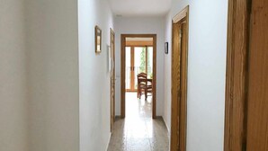 Ein Flur mit einer Holztür rechts, einem Holzrahmen links und einer Tür am Ende, die in einen Raum mit Stuhl und Tisch führt.