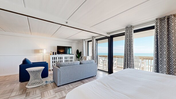 Una habitación de hotel moderna con una cama grande, un sofá, una mesita y un televisor. La habitación tiene un balcón con vistas al mar.