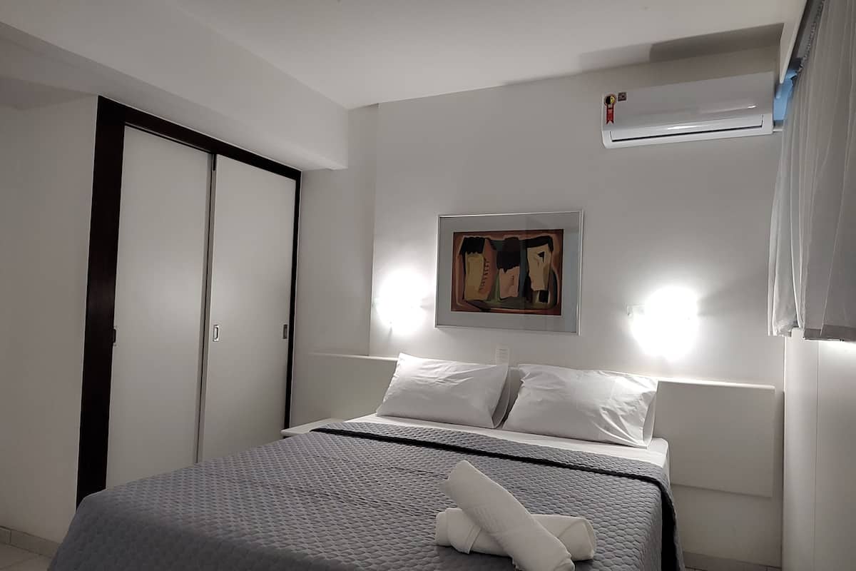 A neatly made bed with a grey quilt, white pillows, and a rolled white towel. There is a framed artwork above the bed and a window with curtains on the right.