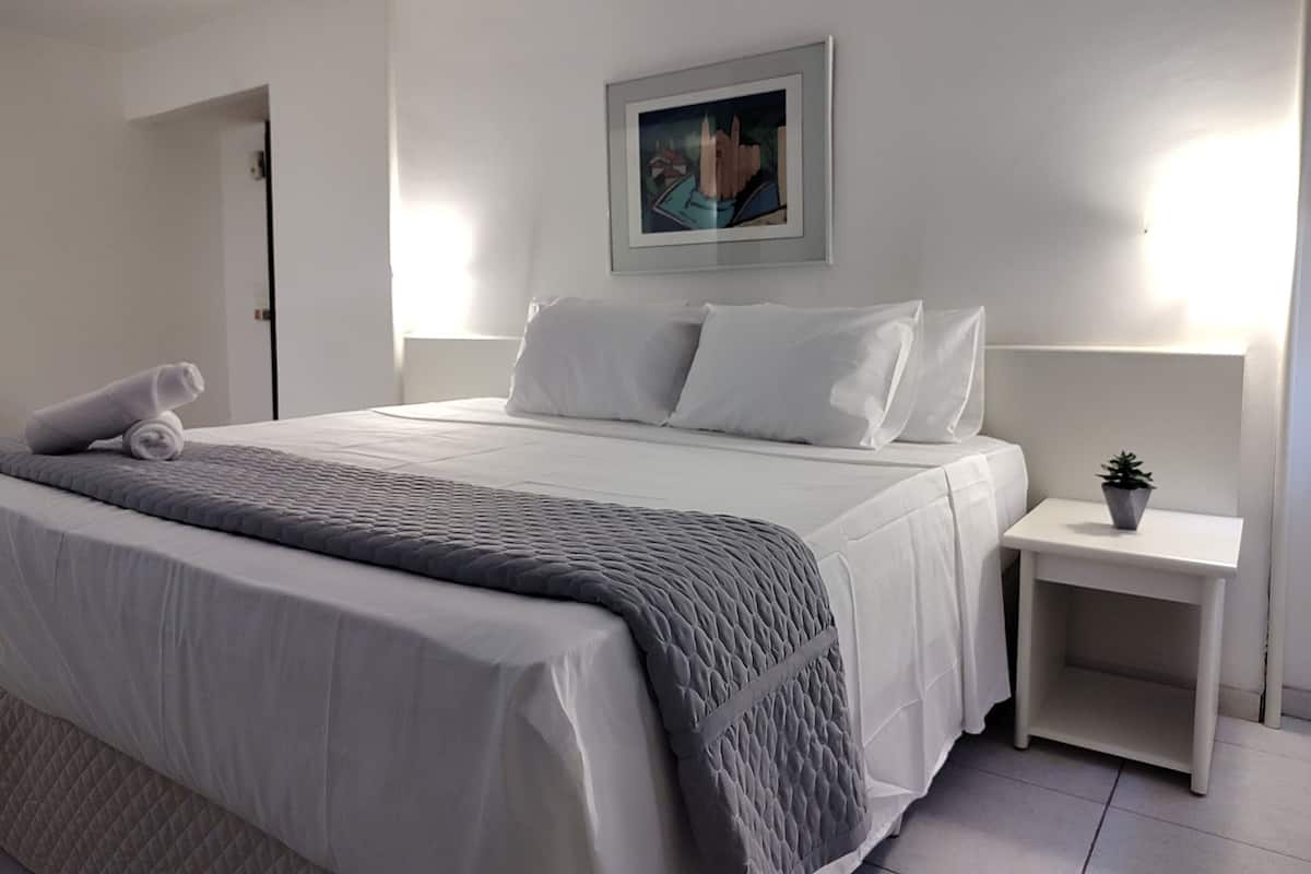 A neatly made bed with a grey quilt, white pillows, and a rolled towel on the side. There is a small bedside table with a plant on it.