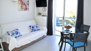 Una sala de estar moderna con un sofá, una mesa de vidrio y sillas. Hay un televisor montado en la pared y una ventana con cortinas.