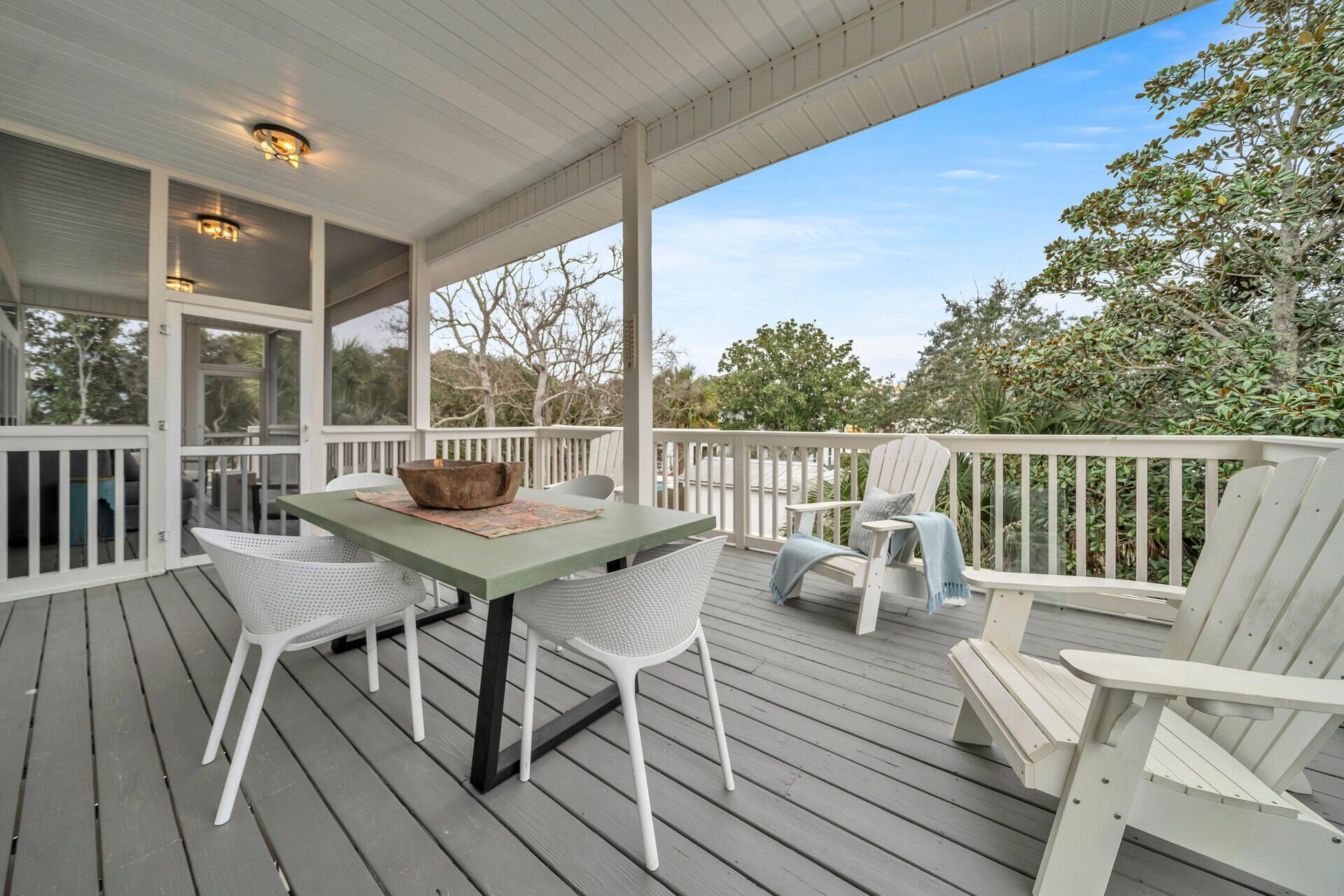 A covered outdoor deck with a table and chairs, surrounded by a white railing and trees in the background.