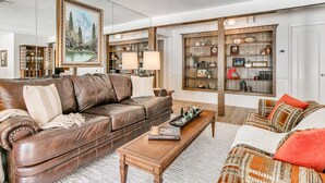 Ein Wohnzimmer mit braunem Ledersofa, einem gemusterten Sessel, einem Holztisch und einem Bücherregal voller Dekoration.