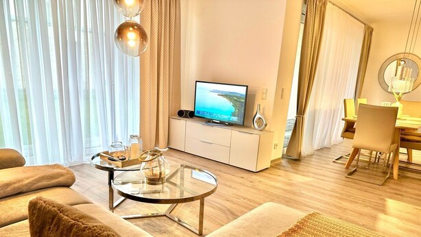 A modern living room with a sofa, a glass coffee table, a TV, and a dining area with a chair and table. - 2802 Wohnung 2 - 2802 - Strandwiese 14 - Wohnung 2 (Dahme)