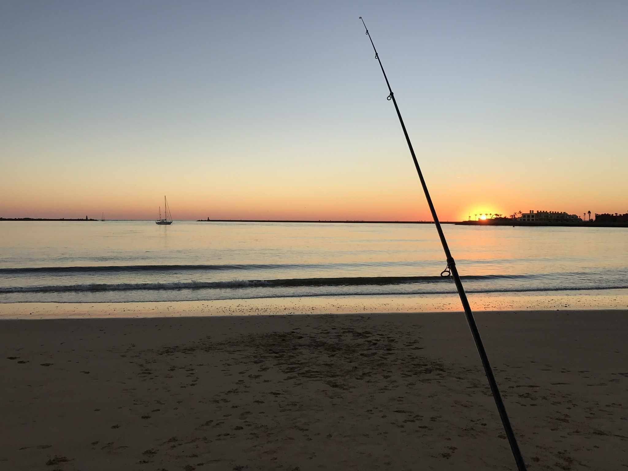 Cena de praia com uma vara de pesca em primeiro plano, mar calmo e um pôr do sol ao fundo.