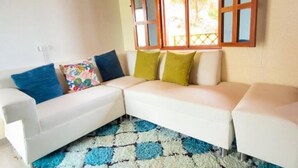 Un canapé d’angle blanc agrémenté de coussins colorés, un tapis à motifs et une fenêtre avec des cadres en bois.