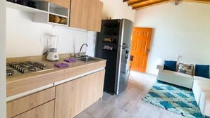 La cuisine est équipée de meubles en bois, d’une cuisinière à gaz, d’un mixeur et d’un réfrigérateur. Le salon comprend un canapé blanc et un tapis bleu.