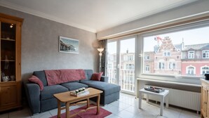 A modern living room with a grey sofa, a wooden coffee table, and a view of red brick buildings through large windows. - Centrally located by Interhome (Blankenberge)