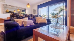 Uma sala de estar moderna com um sofá estampado escuro, duas luminárias pendentes, uma mesa de centro de vidro e uma grande janela com vista para o exterior.