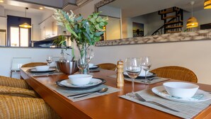 Uma área de refeições com uma mesa de madeira comprida posta para dois, rodeada por cadeiras de vime. Há um vaso com plantas verdes na mesa, e também dois copos de vinho e um saleiro e pimenteiro.