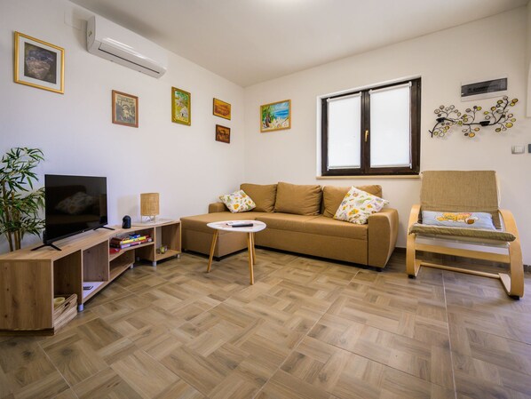 Sala de estar con sofá, televisor, una mesita y una silla. Se observan adornos en la pared y una ventana.